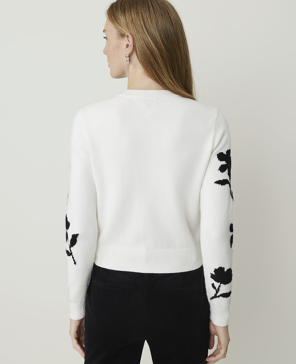 A woman in a white sweater with black floral accents on the sleeves stands against a plain white background.