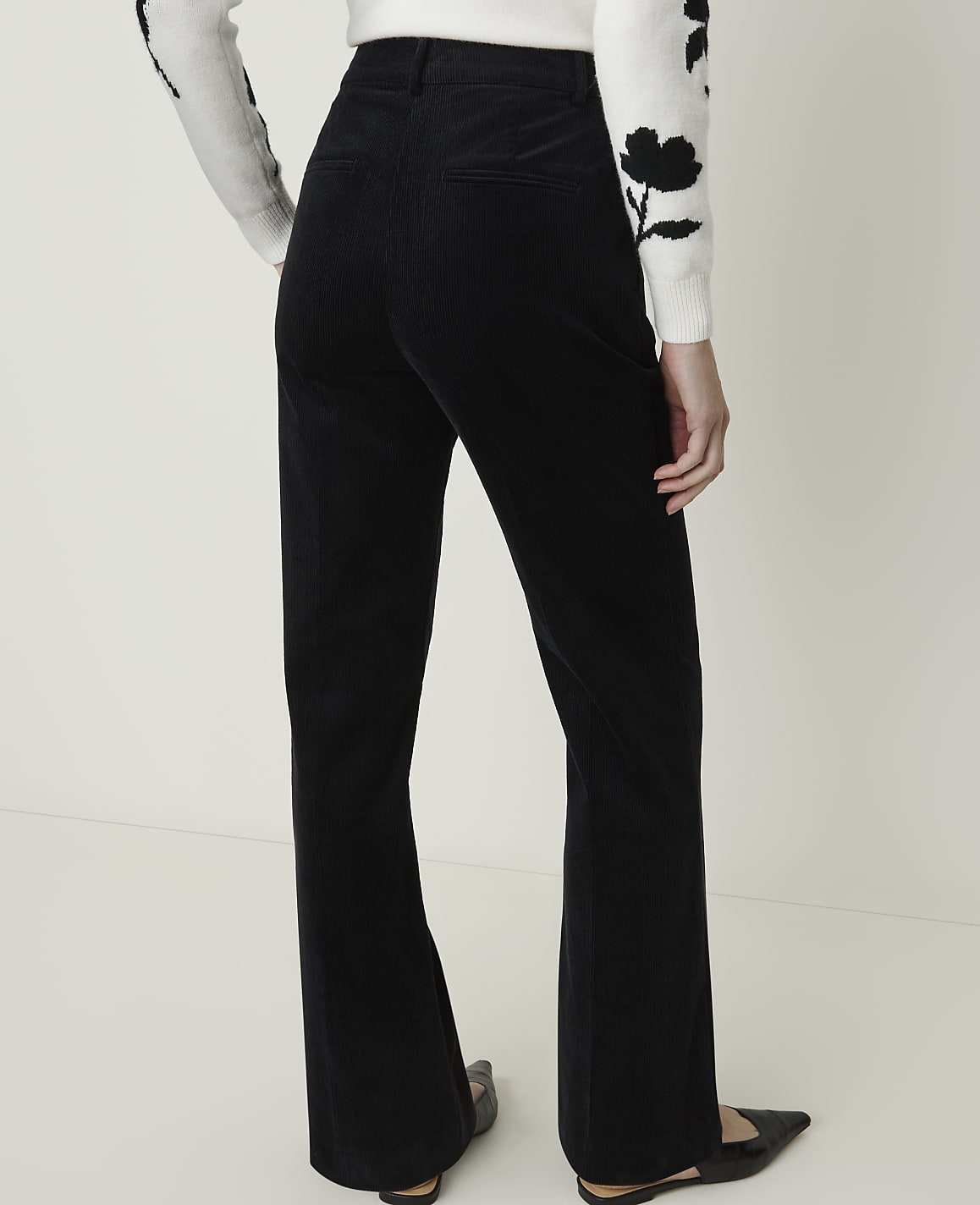 A person wearing black corduroy flared pants and a white sweater with floral accents stands against a plain white background.
