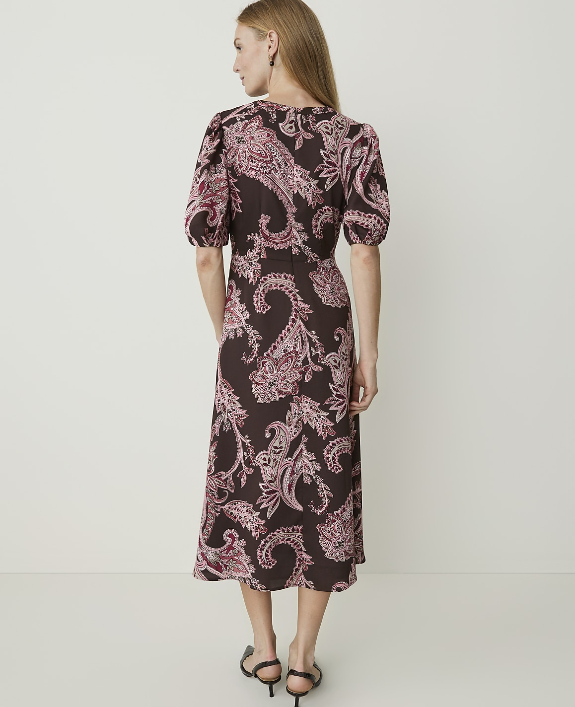 A woman in a brown paisley dress stands against a plain white background.