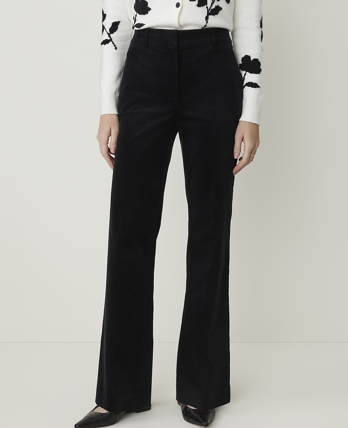 A person stands in black corduroy flared pants and a white floral sweater against a plain white background.