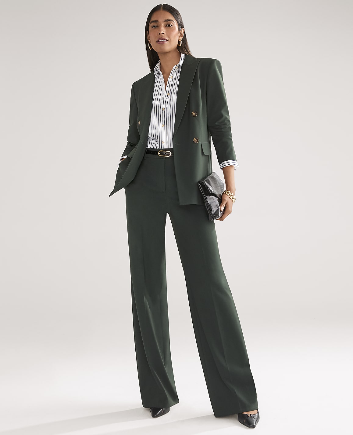 A woman in a dark green pantsuit stands against a plain, light gray background, holding a black clutch.