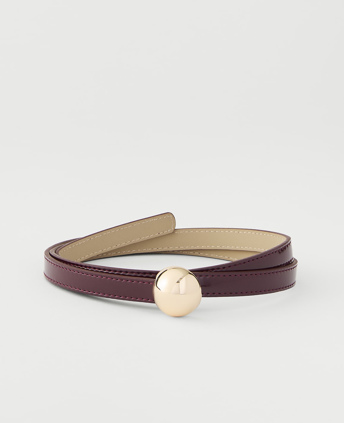 A thin, glossy burgundy belt with a gold spherical buckle is coiled on a plain white background.