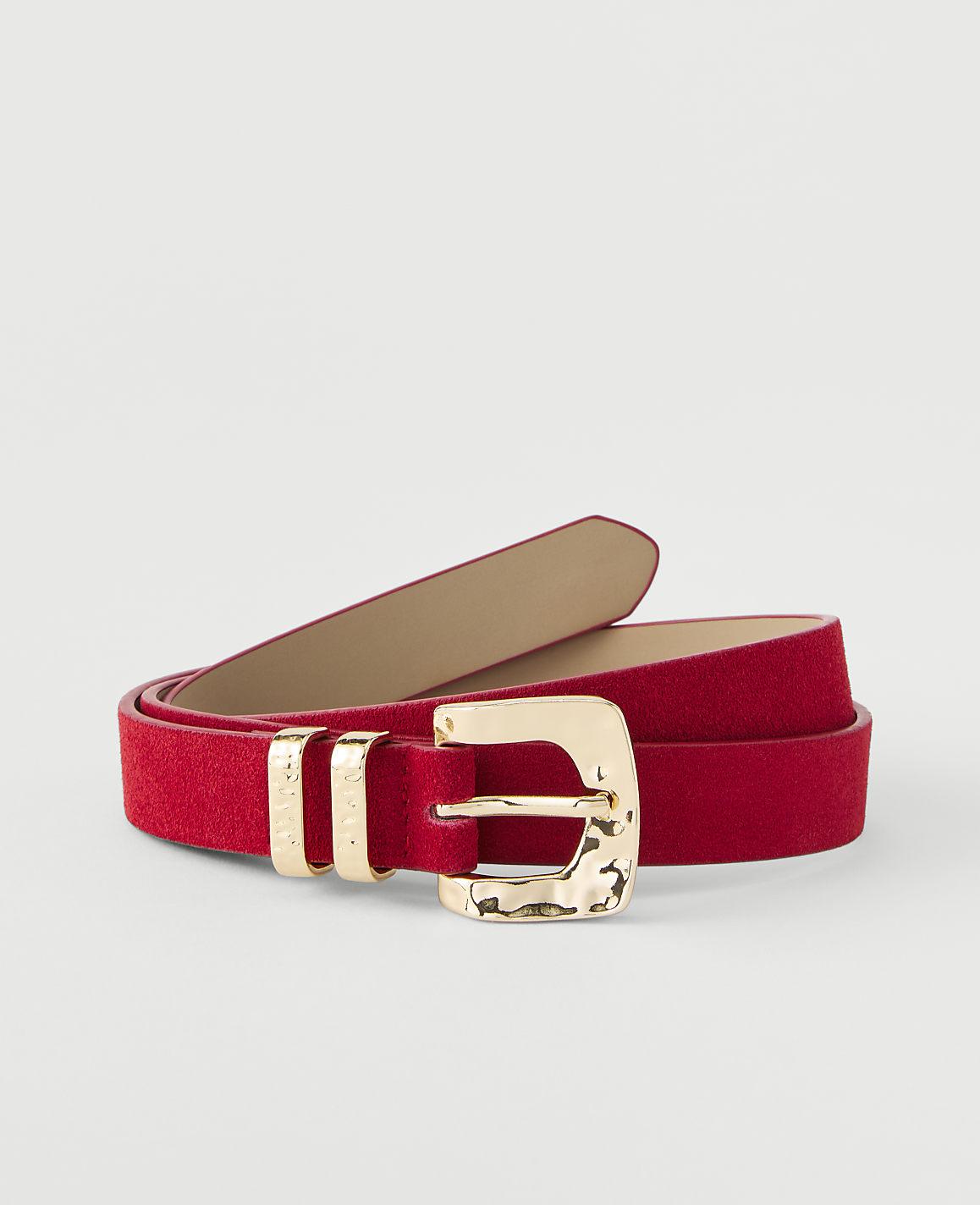 A red suede belt with a hammered gold buckle lies coiled on a plain white background.