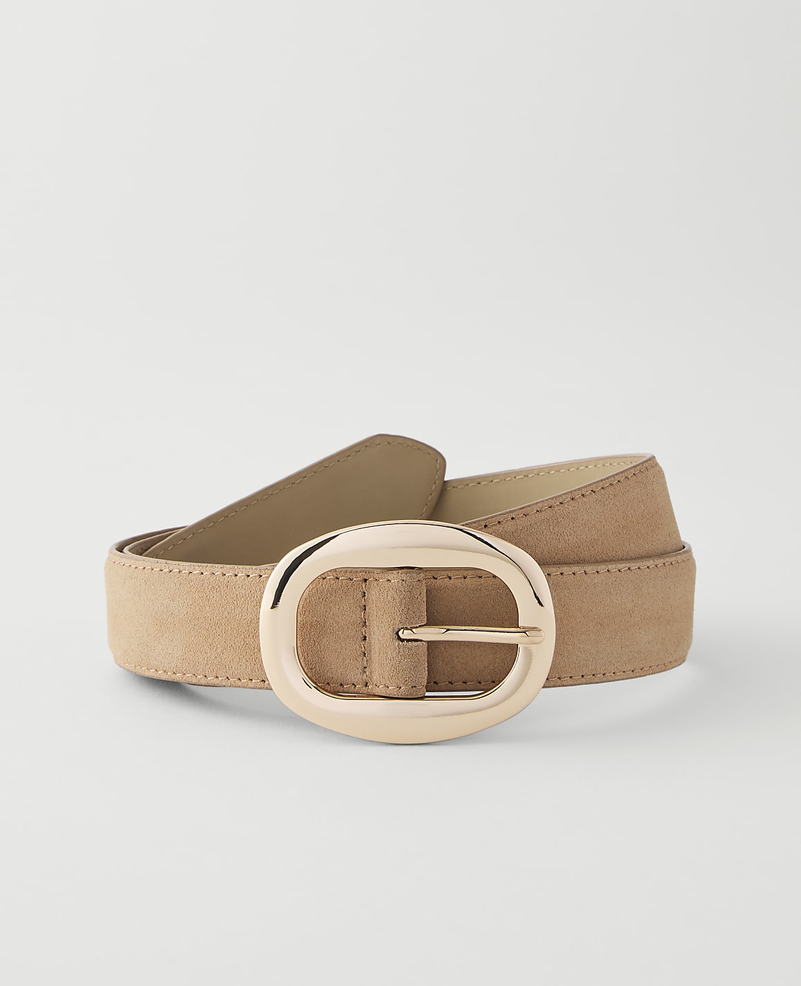 A tan suede belt with a large, oval, rose gold buckle lies on a plain white background.