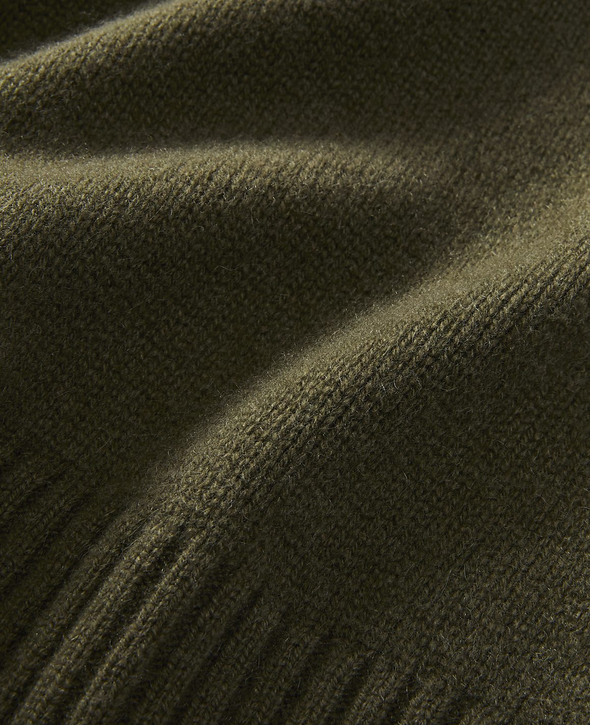 A close-up of a textured olive green knitted fabric fills the frame, with soft folds and a ribbed edge visible in the foreground and a smooth, slightly blurred expanse in the background.
