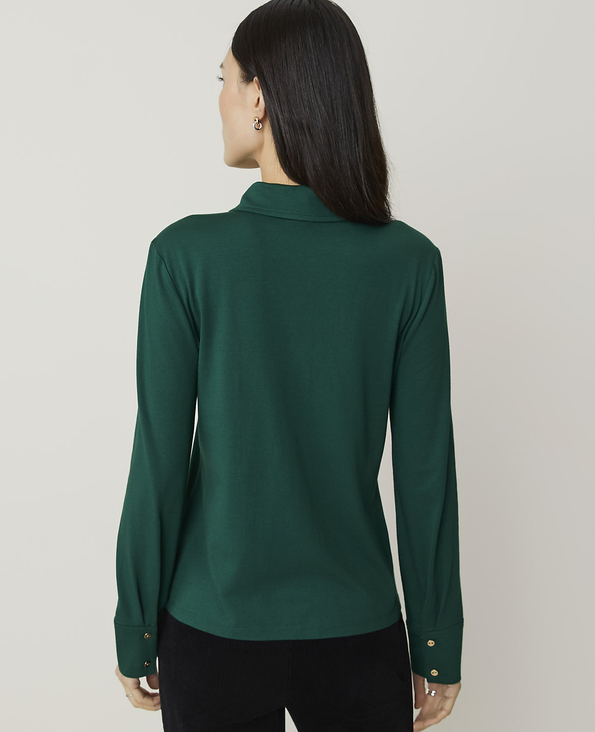 A woman in a green collared shirt stands against a plain, light gray background.