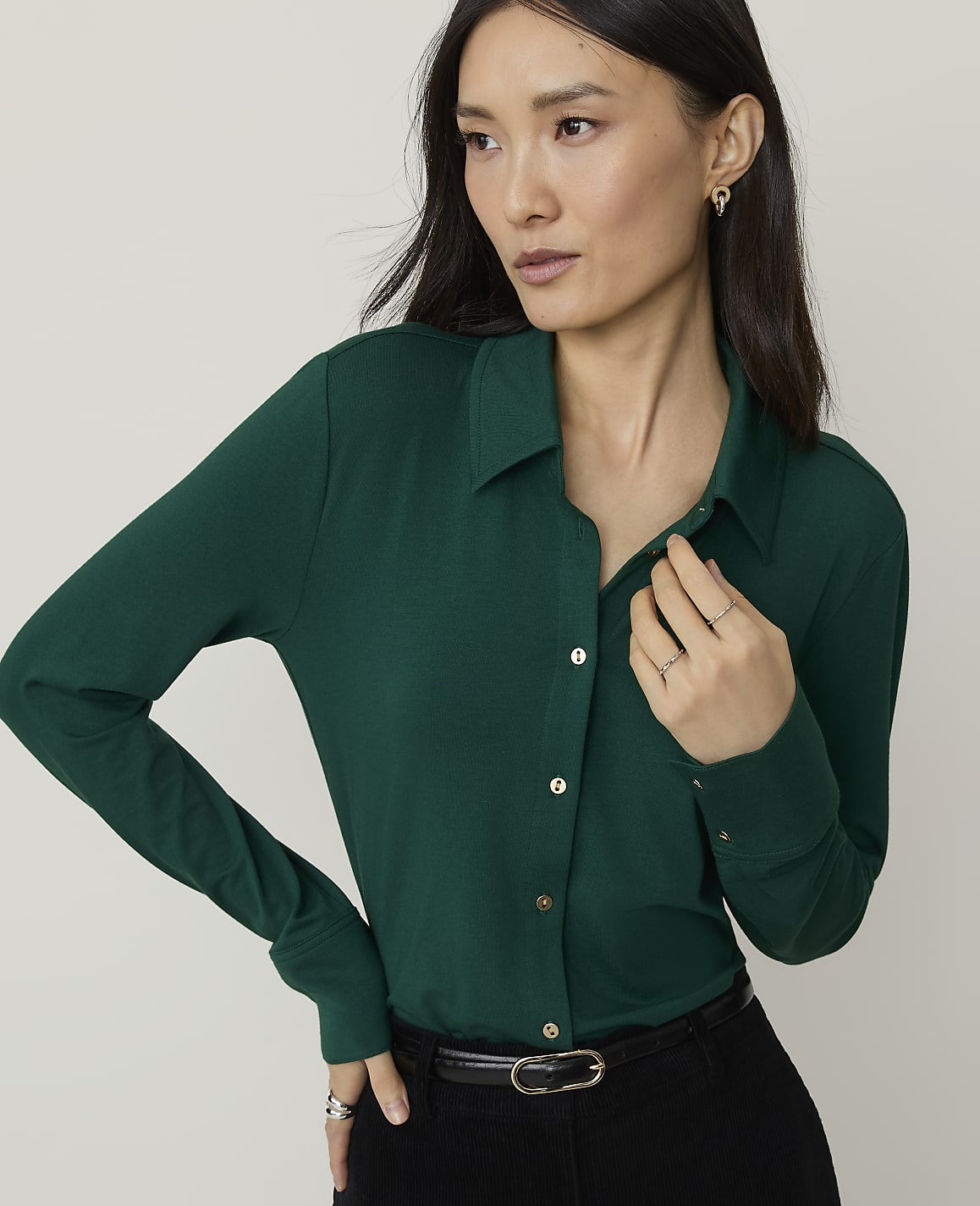 A woman in a green collared shirt stands against a plain, light gray background.