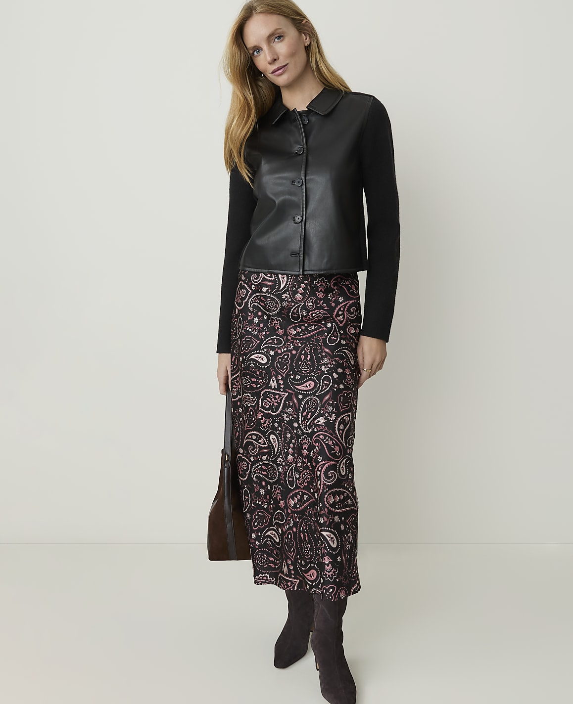A woman in a black leather jacket and a patterned skirt stands against a plain white background, holding a brown handbag.