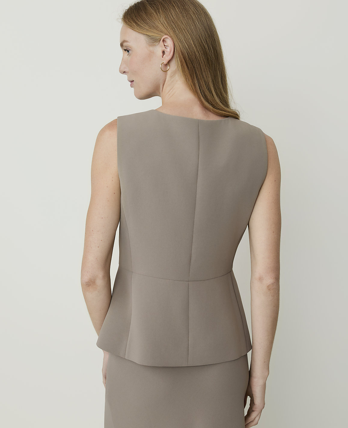 A woman in a taupe peplum top and skirt stands against a plain, light background.