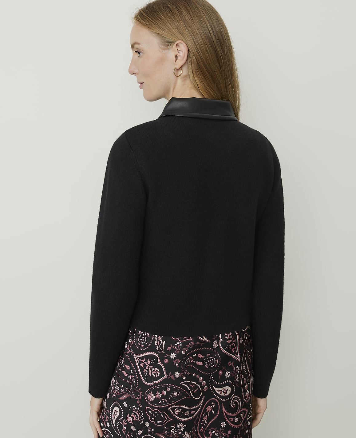 A woman in a black sweater with a leather collar and a patterned skirt stands against a plain, light-colored background.