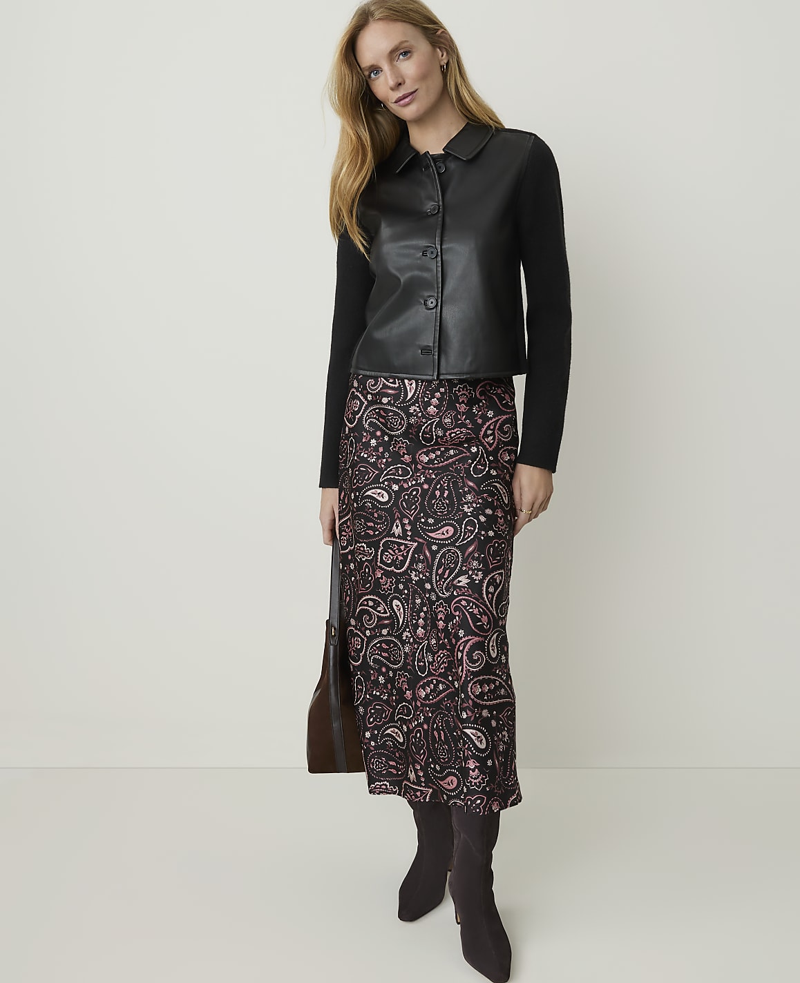 A woman in a black leather-look jacket and a patterned skirt stands against a plain, light-colored background.