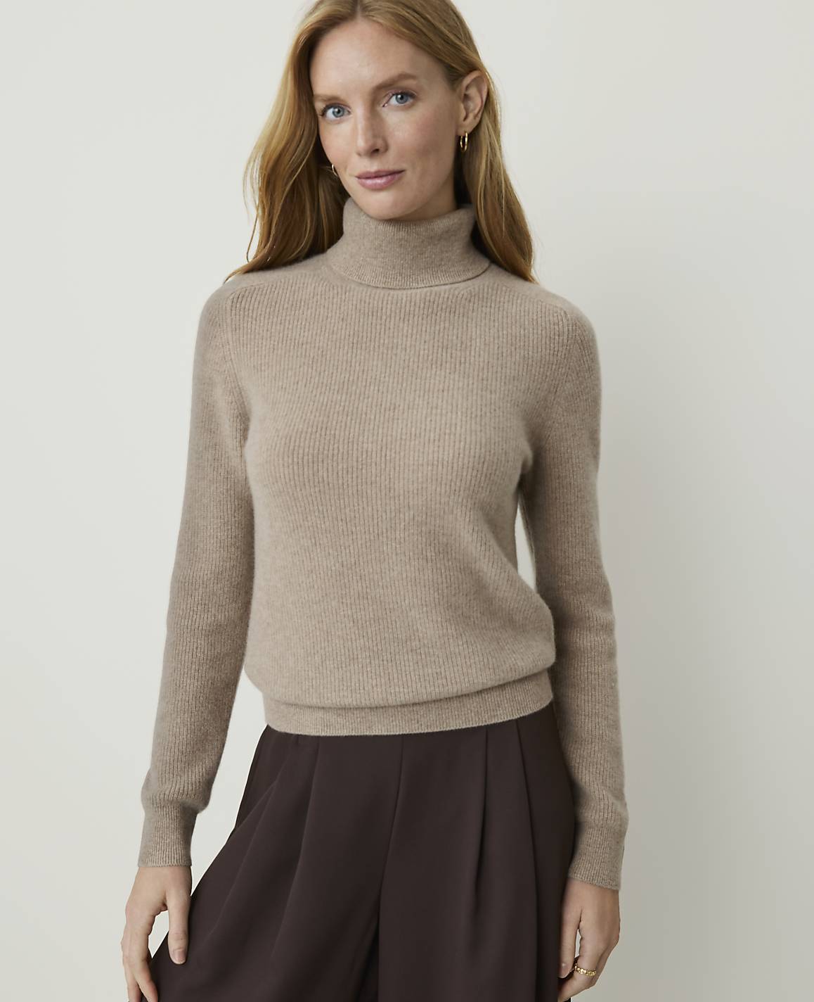 A woman in a beige turtleneck sweater and brown pants stands against a plain, light background.