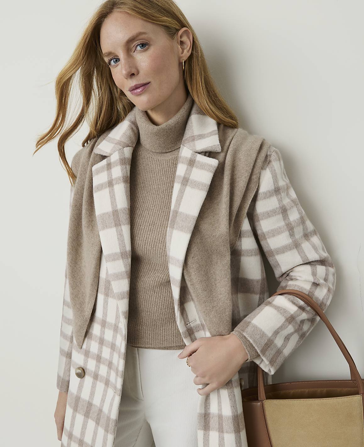 A woman in a beige turtleneck and a plaid coat stands against a plain, light-colored wall, holding a brown tote bag.