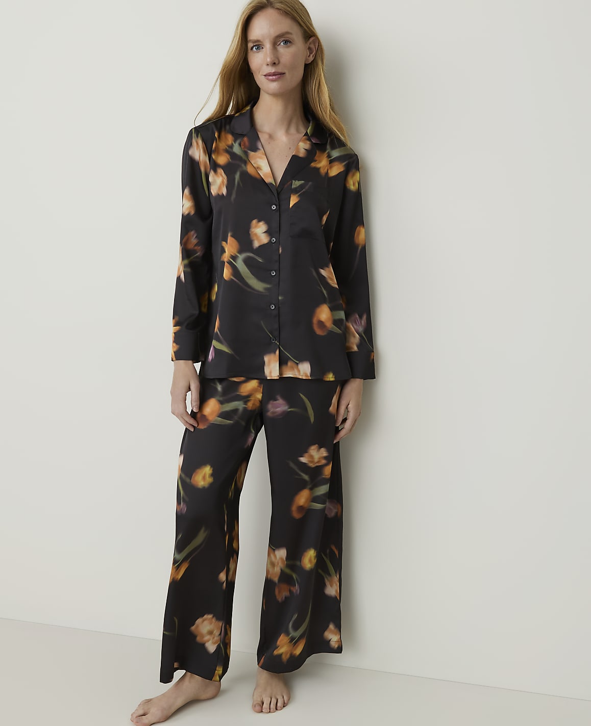 A woman stands in a black floral pajama set against a plain, light-colored wall.