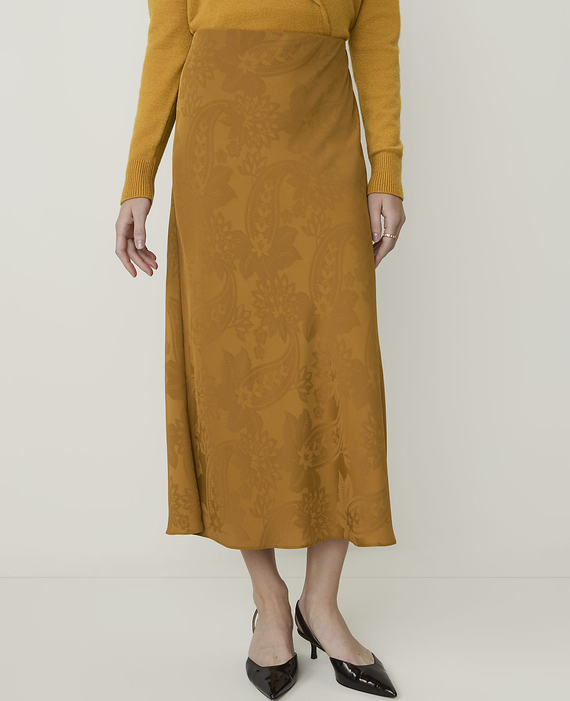 A woman in a mustard yellow sweater and a patterned midi skirt stands against a plain, light background.