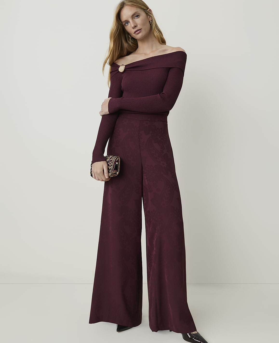 A woman in a burgundy off-the-shoulder jumpsuit and patterned clutch stands against a plain, light-colored background.