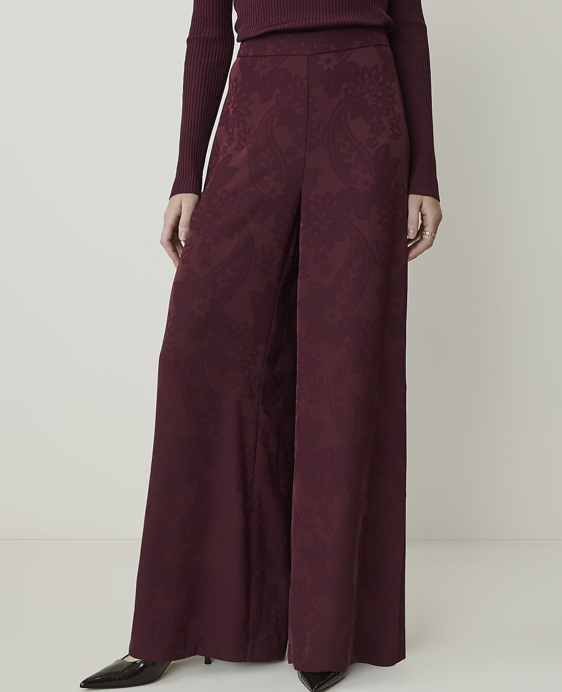 A person stands in wide-leg burgundy patterned pants and a ribbed sweater against a plain white background.