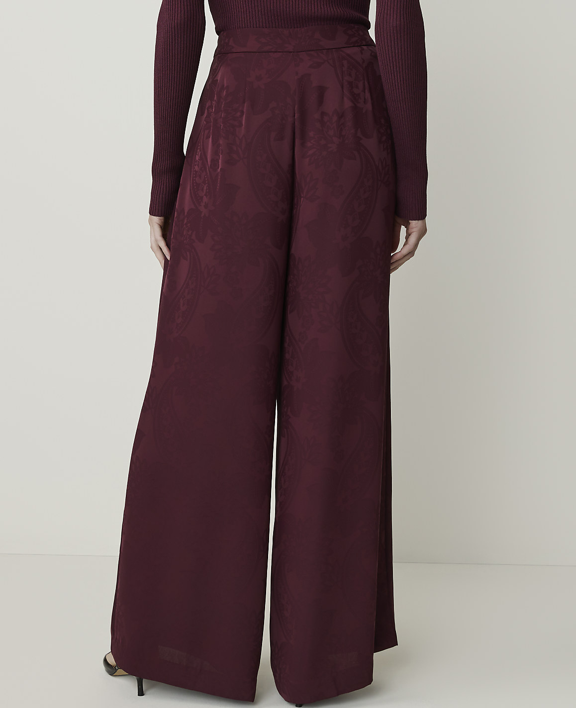 A person stands in wide-leg burgundy pants with a paisley pattern in the foreground, against a plain white background.