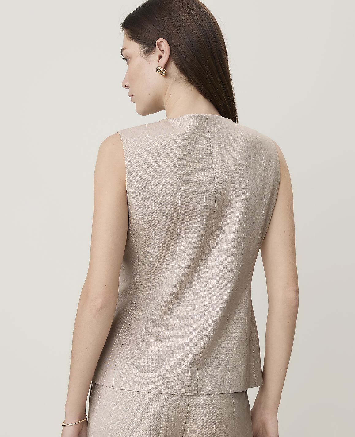 A woman in a beige plaid vest and pants stands against a plain, light-colored background.