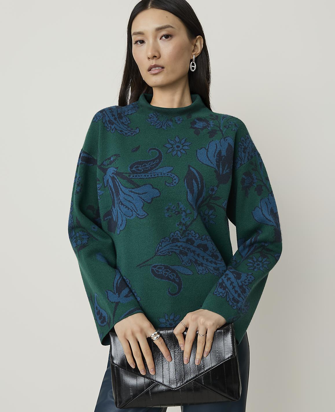 A woman in a green patterned sweater holds a black clutch in front of a plain background.