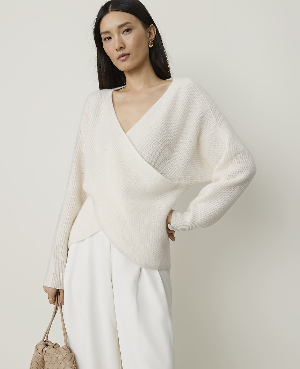 A woman in a cream wrap sweater and white pants stands against a plain, light background, holding a woven bag.