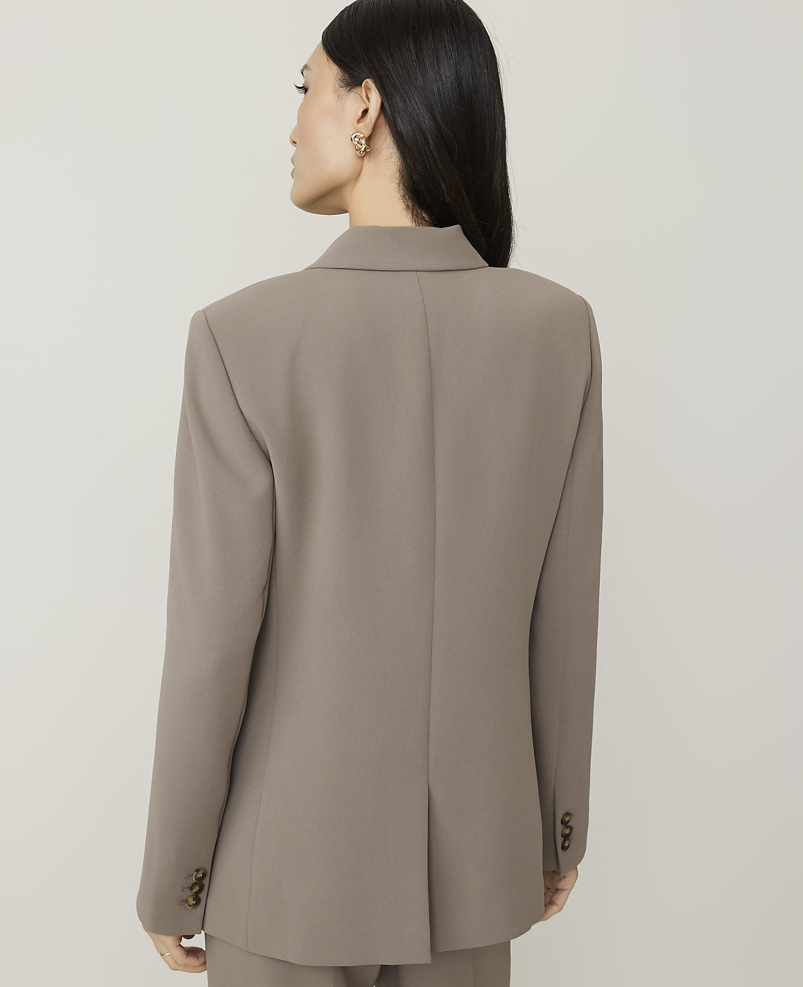A woman in a taupe blazer stands against a plain, light-colored background.