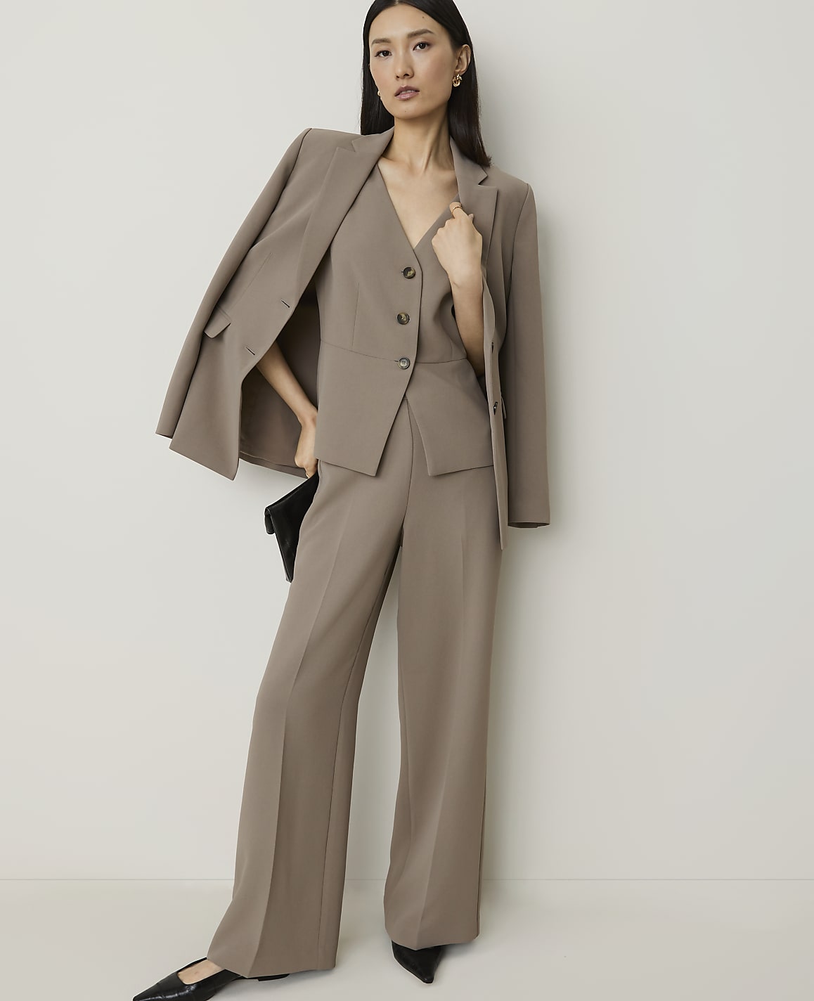 A woman in a taupe pantsuit with a blazer draped over her shoulder poses against a plain white background.