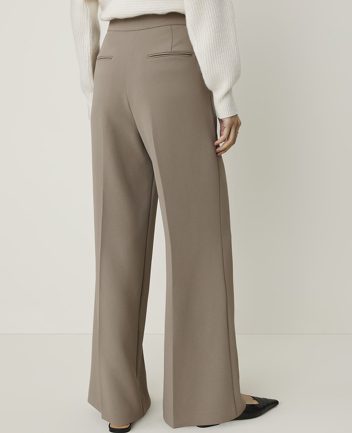 A person wearing wide-leg taupe pants and a white sweater stands against a plain white background.