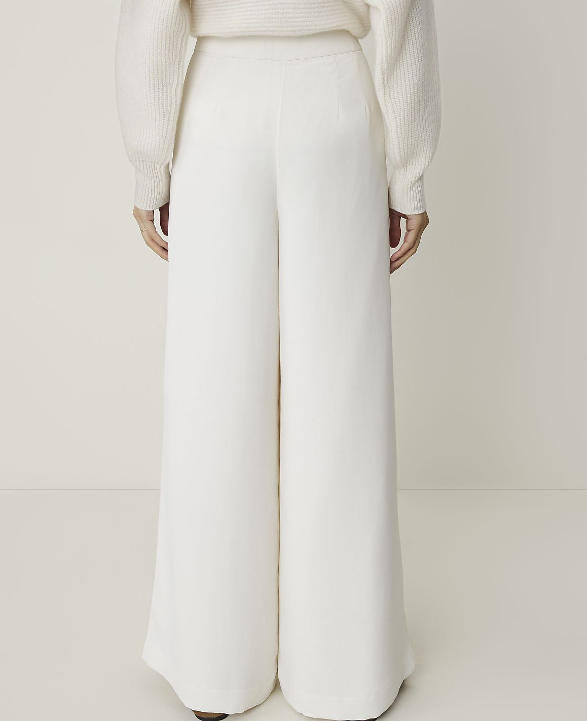 A person wearing white wide-leg pants and a cream sweater stands against a plain white background.