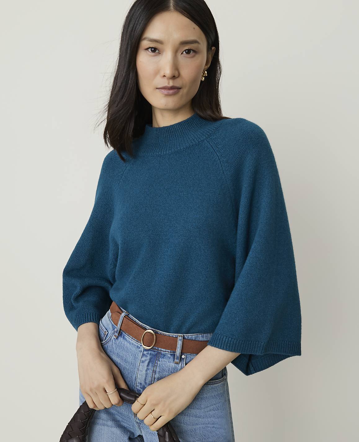 A woman in a teal sweater and jeans stands against a plain background.