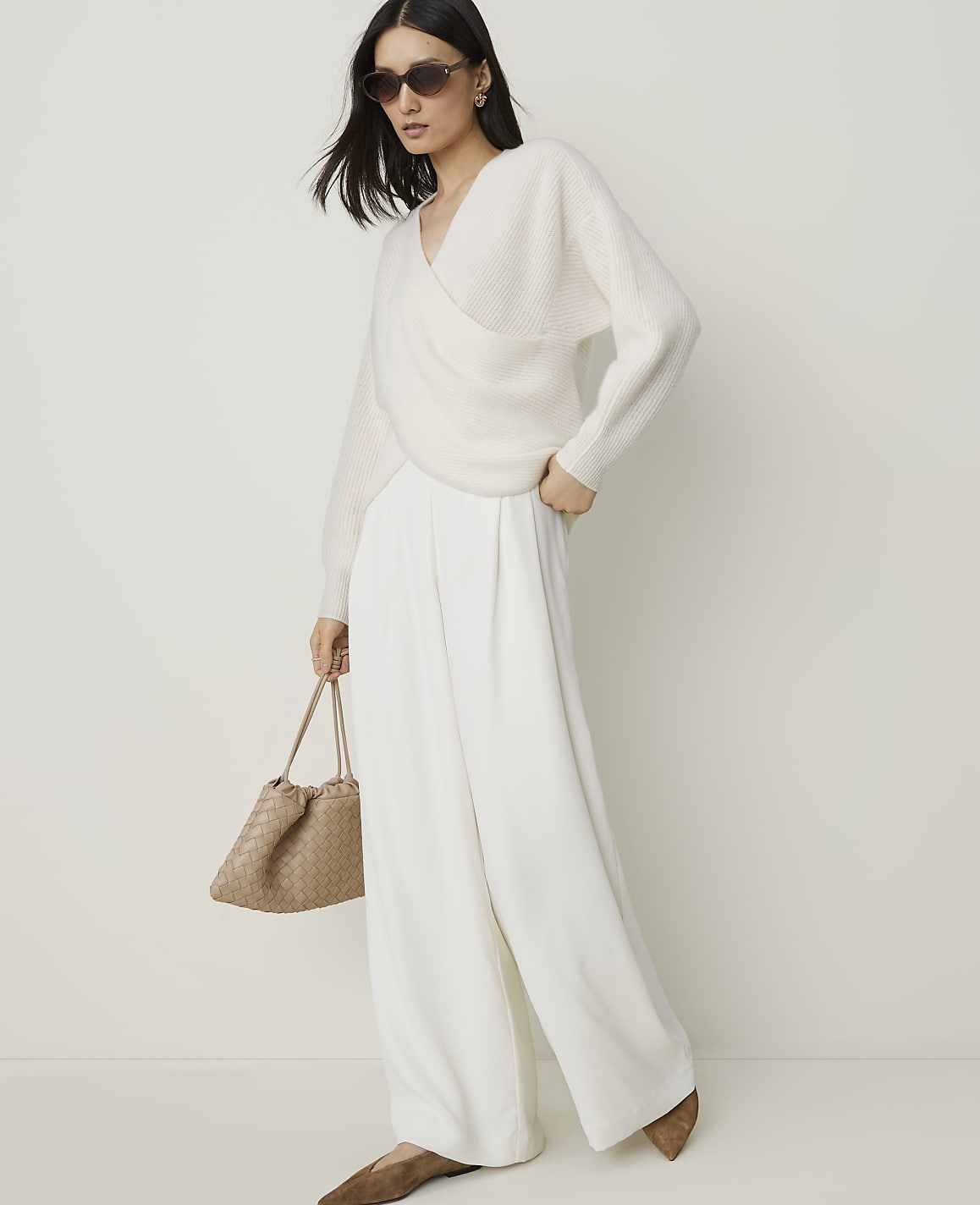A woman in a cream wrap sweater and wide-leg pants poses against a plain, light background, holding a woven handbag.