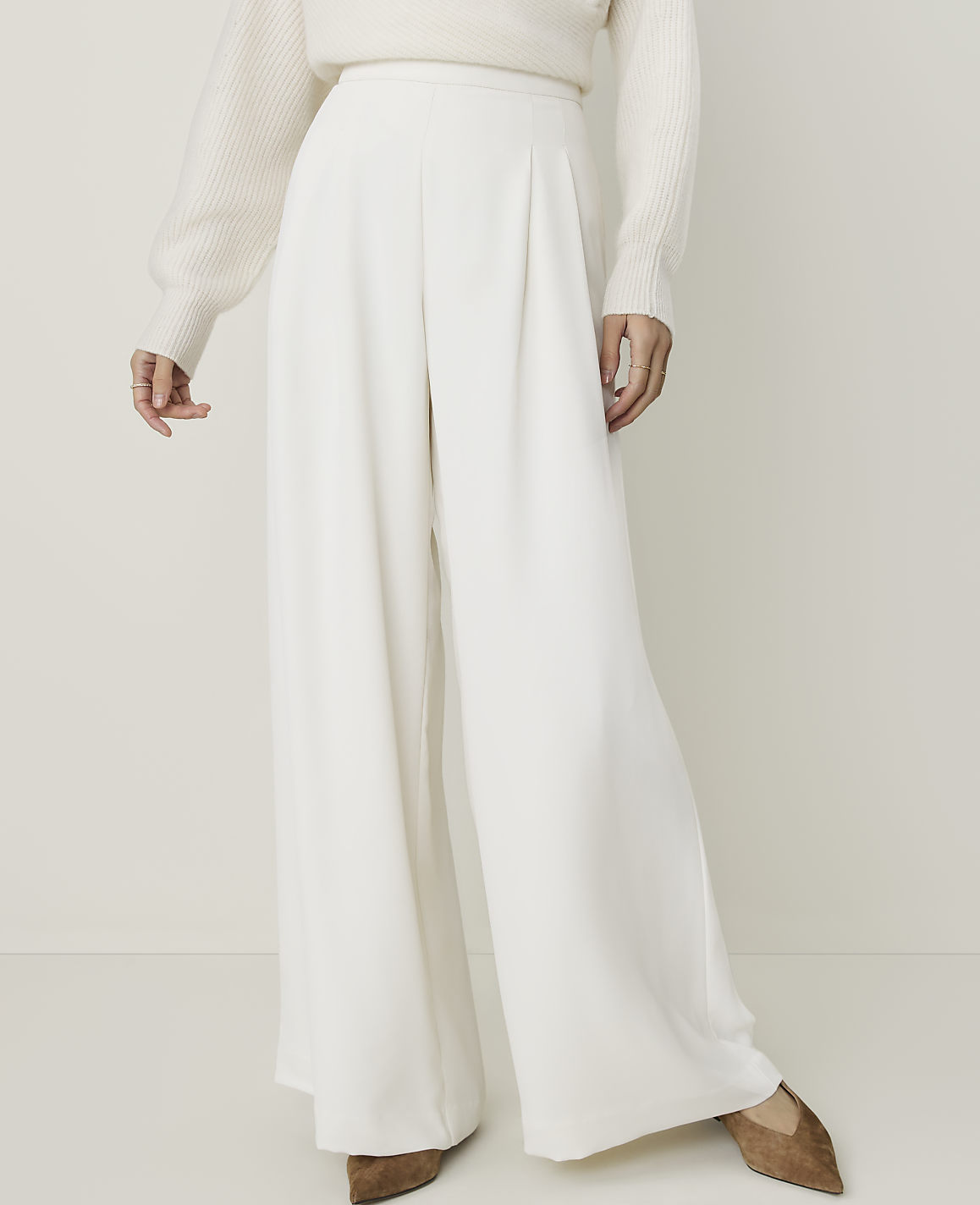 A person in a cream sweater and white wide-leg pants stands against a plain, light background.