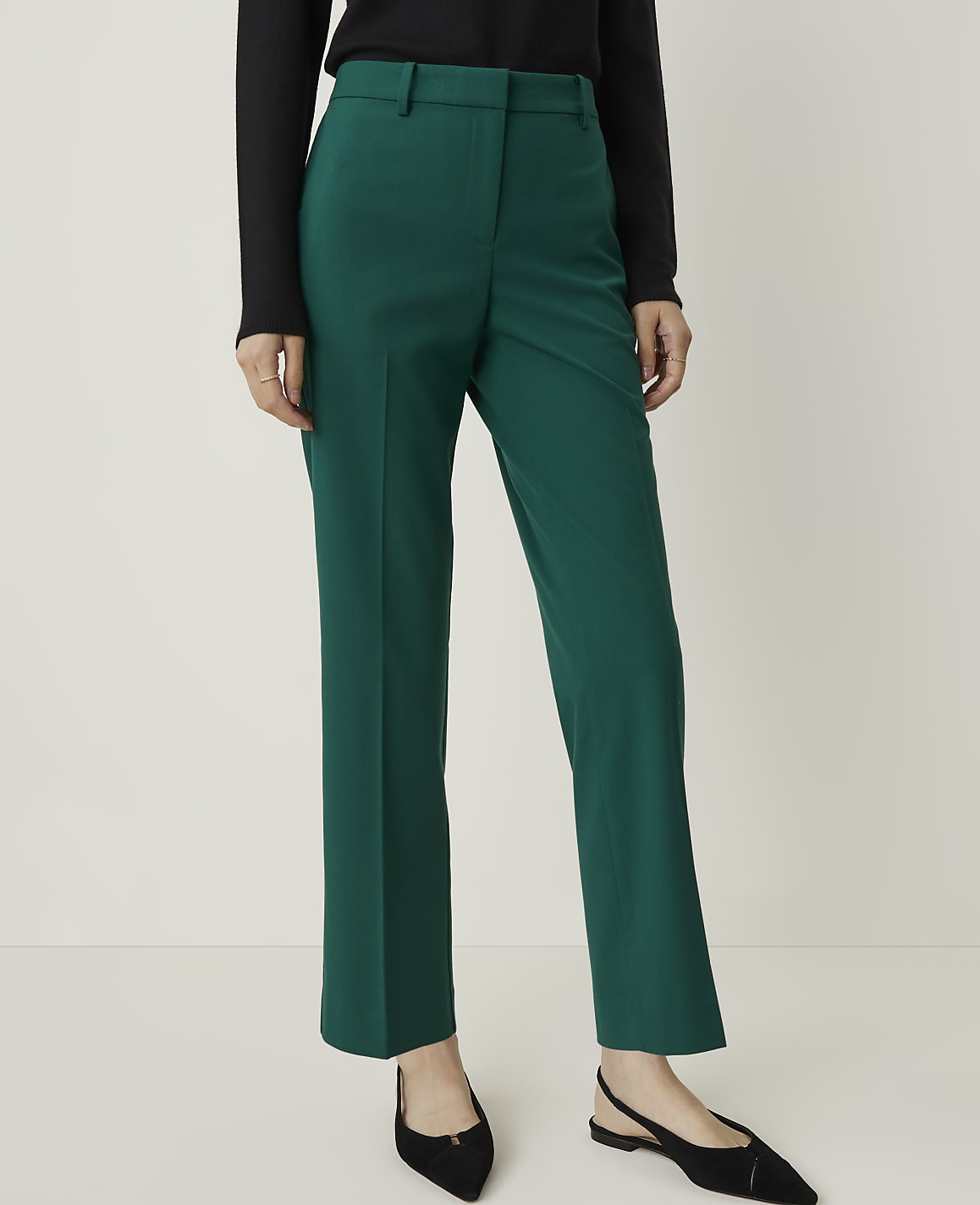 A person wearing emerald green trousers and black pointed flats stands against a plain, light gray background.
