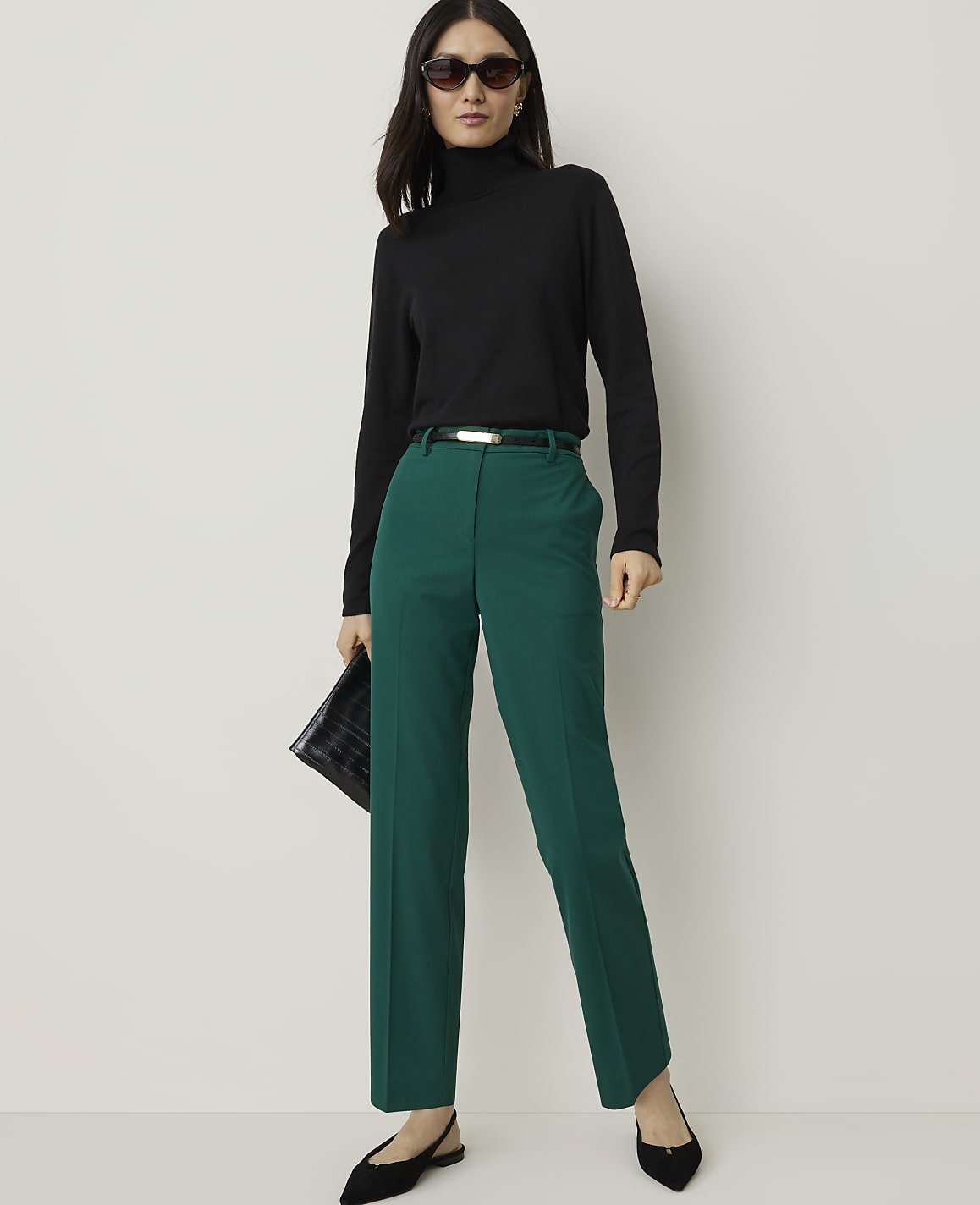 A woman in a black turtleneck and green pants stands against a plain white background, holding a black clutch.