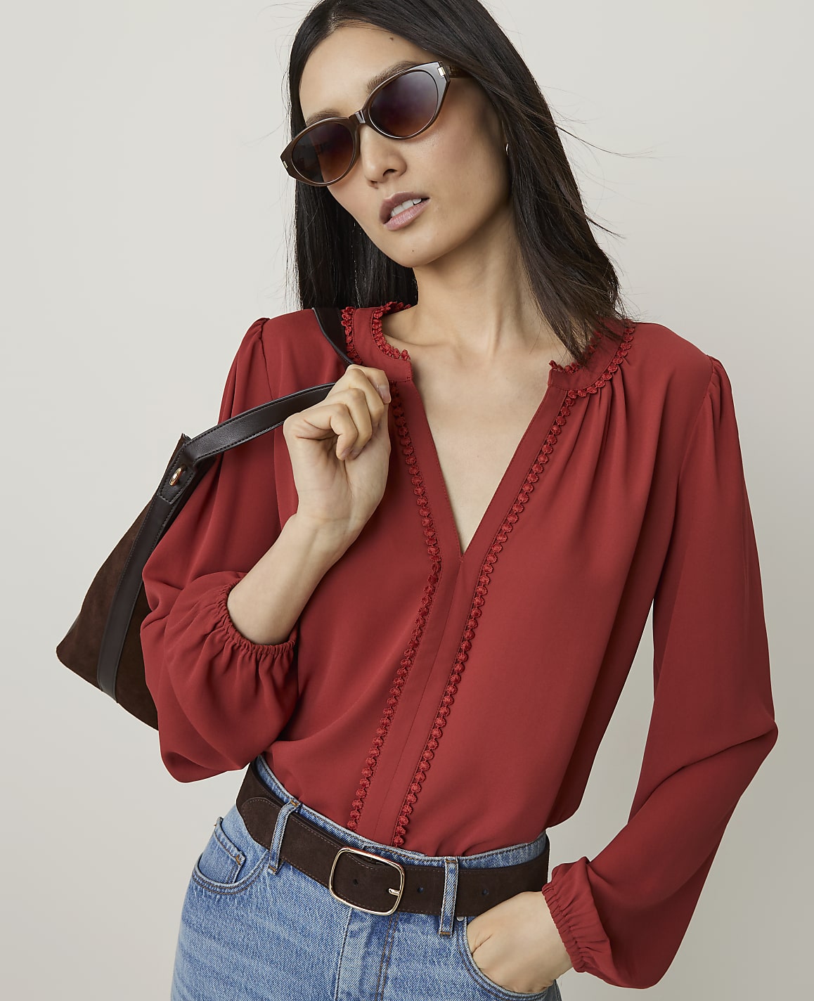 A woman in a red blouse and jeans, wearing sunglasses and carrying a brown bag, stands against a plain background.
