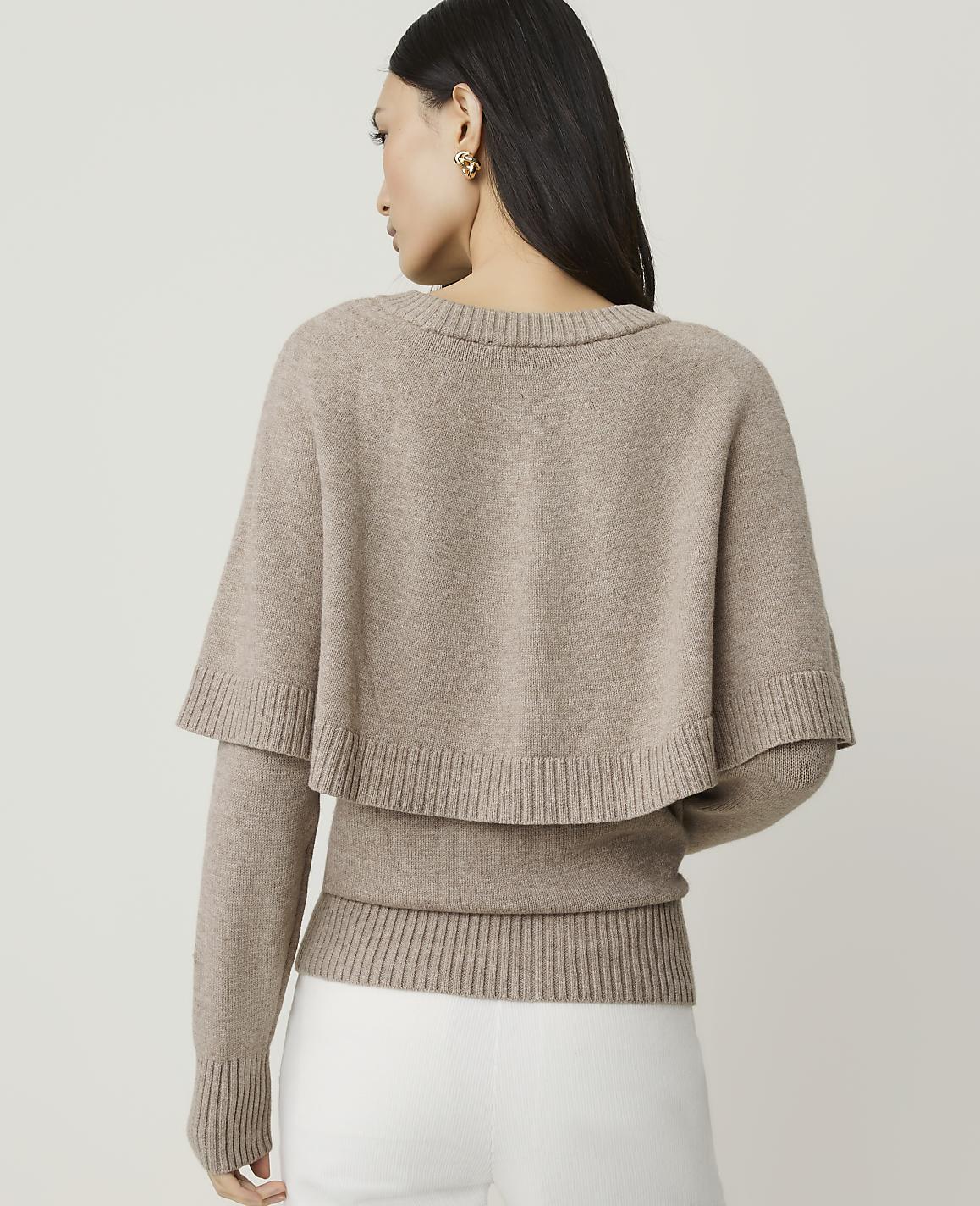A woman in a beige sweater with a capelet stands against a plain, light background.