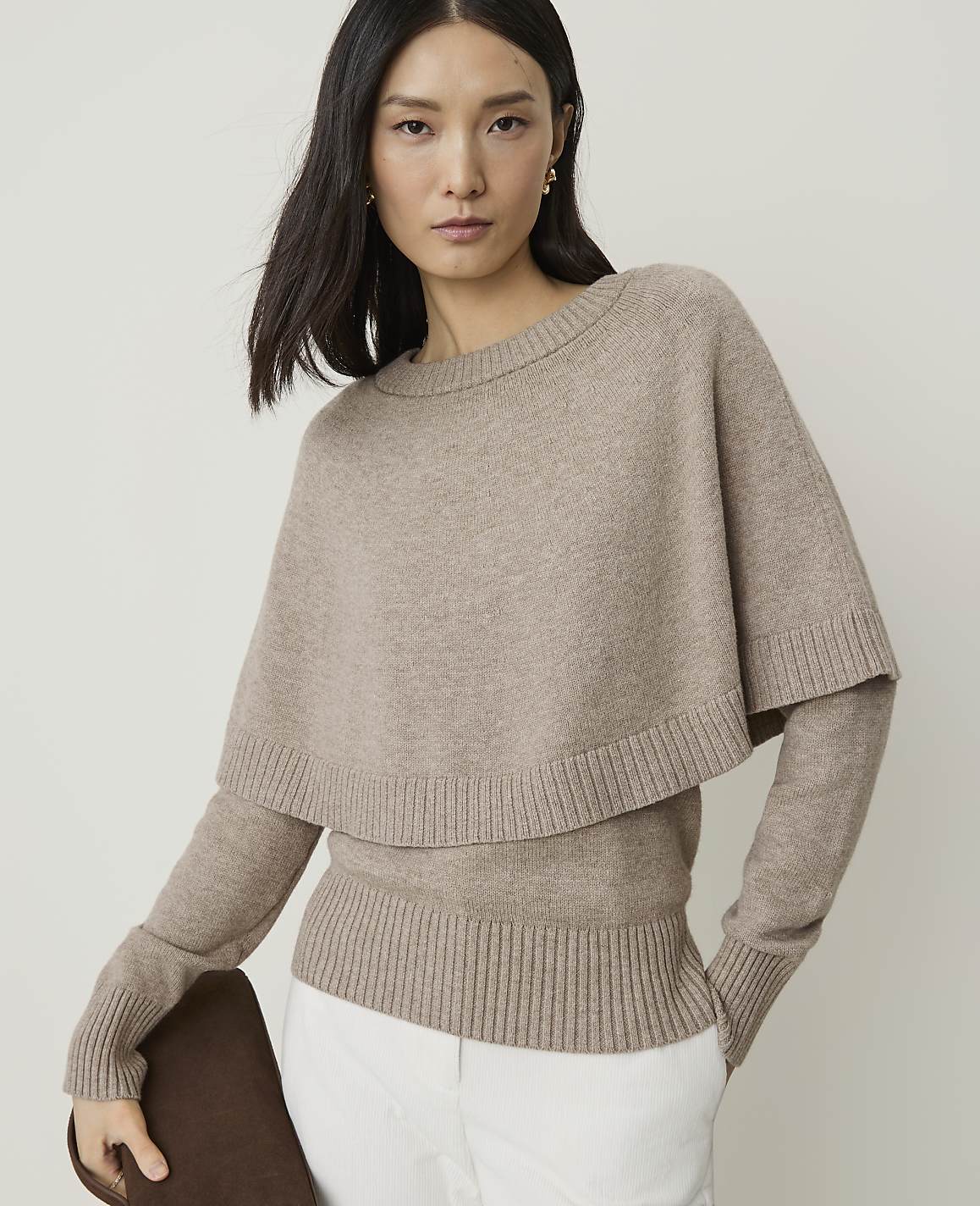 A woman in a beige sweater with a cape overlay stands against a plain, light background.