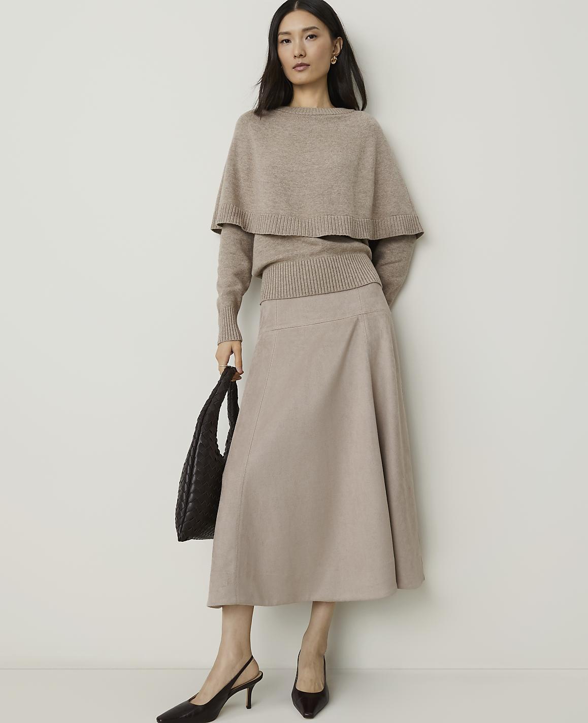 A woman in a beige sweater and skirt stands against a plain white background, holding a dark brown handbag.