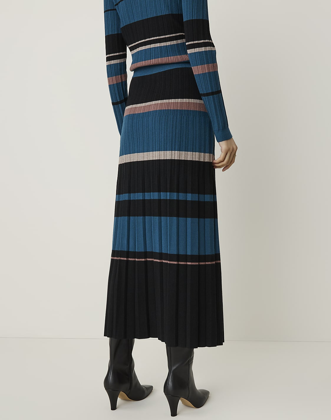 A person stands in black boots, wearing a pleated midi skirt and a matching long-sleeved top with horizontal stripes of teal, black, and pink against a plain, light background.