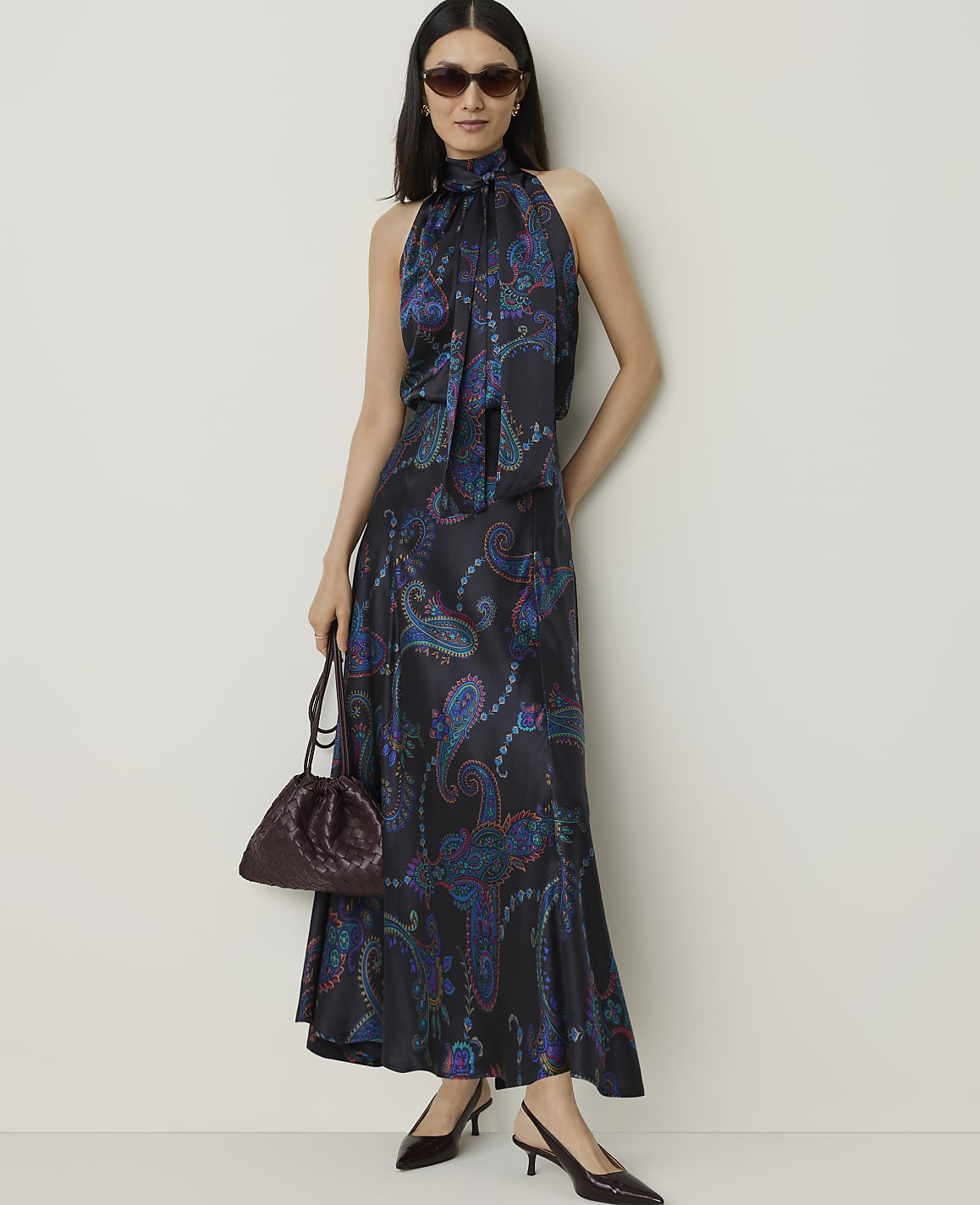 A woman in a patterned halter dress and sunglasses stands with a handbag in the foreground against a plain wall in the background.