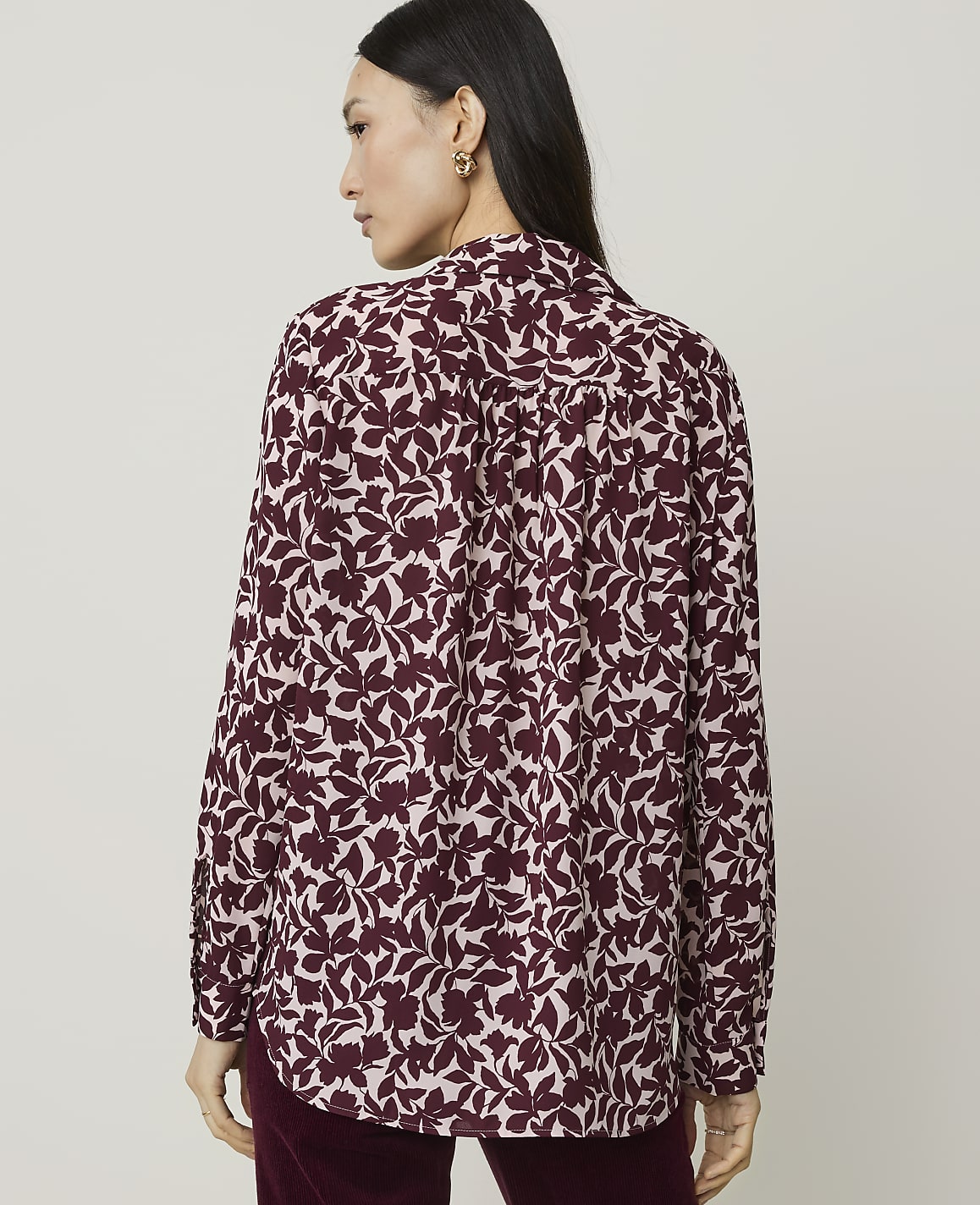A woman in a burgundy floral print blouse stands against a plain, light-colored background.