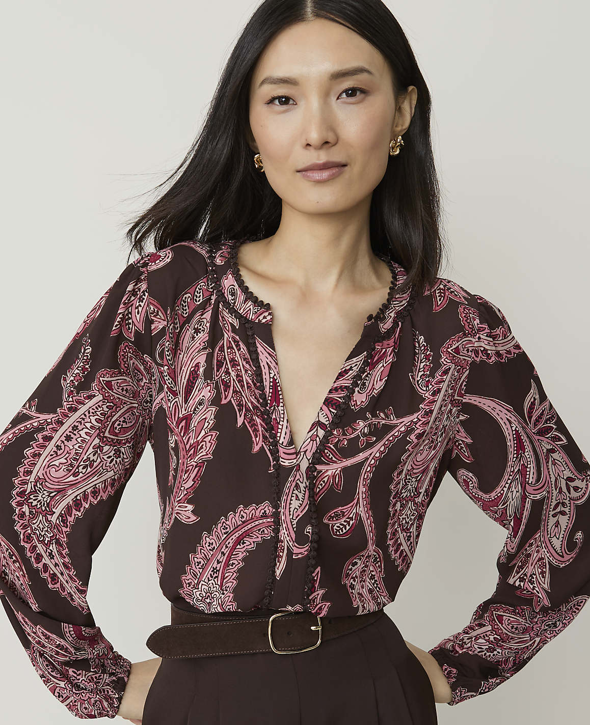 A woman with dark hair and a patterned blouse stands against a plain background.