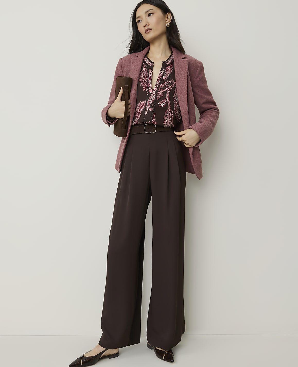 A woman in a pink blazer and brown wide-leg pants stands against a plain, light-colored background, holding a brown clutch.