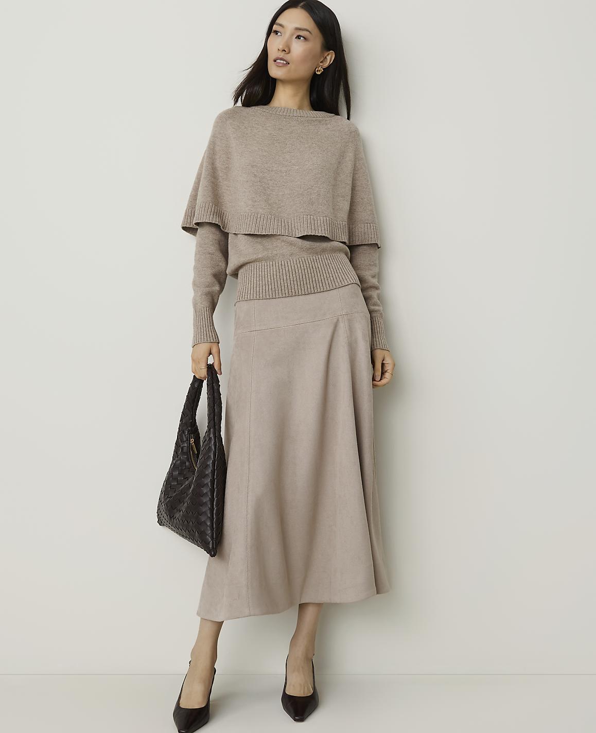 A woman in a beige sweater and skirt stands against a plain white background, holding a dark woven handbag.