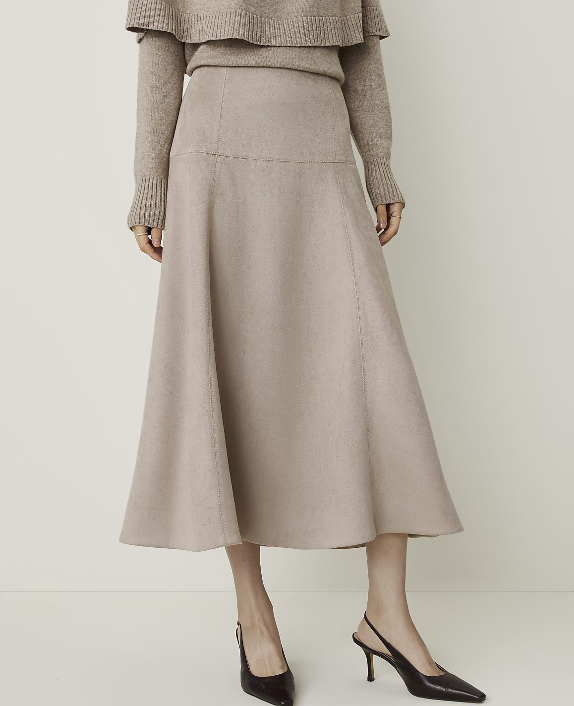 A woman in a beige sweater and a flowing beige skirt stands against a plain, light-colored background, wearing dark slingback heels.
