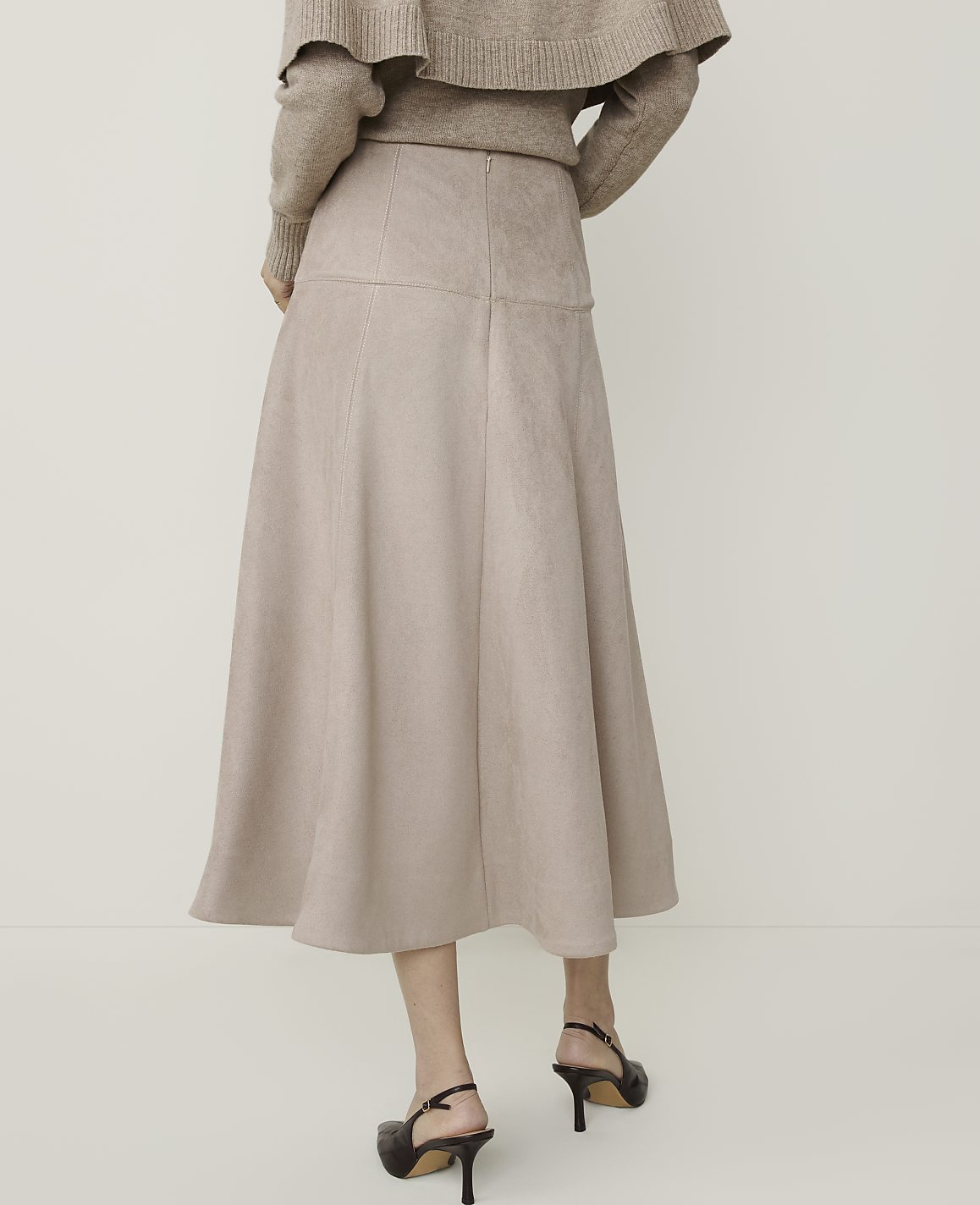 A woman in a beige sweater and a flowing beige skirt stands against a plain white background.