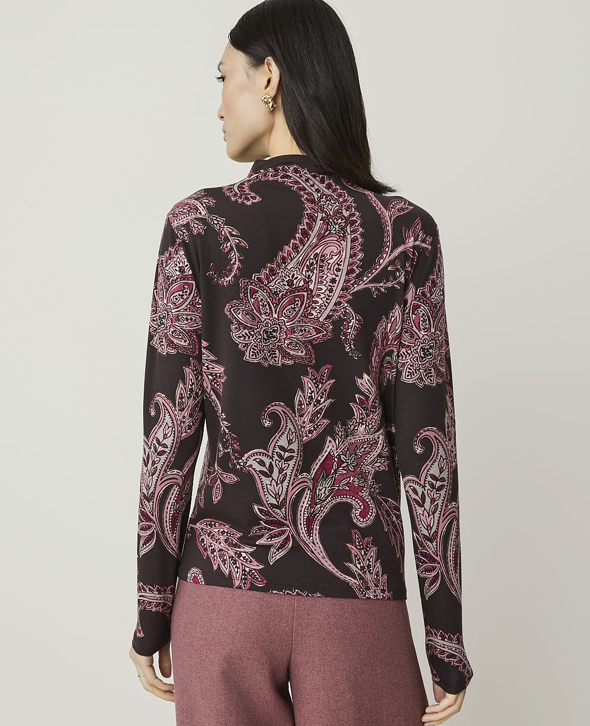 A woman in a dark, paisley-patterned top and pink pants stands against a plain, light gray background.