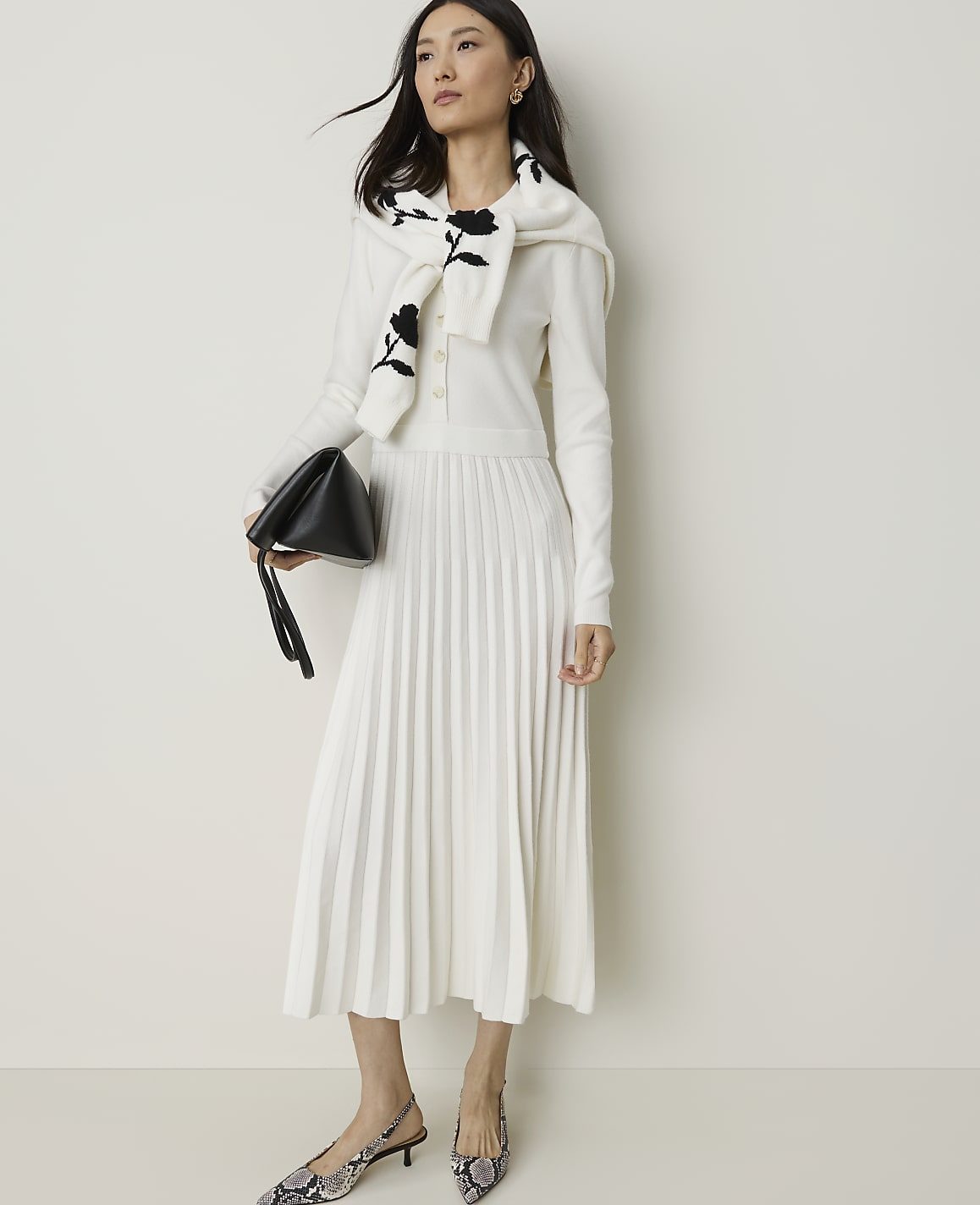 A woman in a white pleated dress and a floral-patterned sweater draped over her shoulders holds a black clutch while standing against a plain white background.
