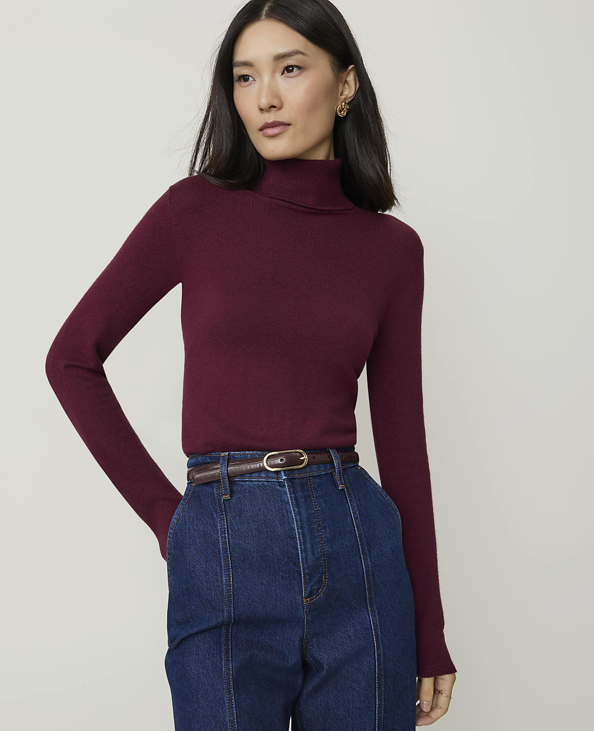 A woman in a burgundy turtleneck and blue jeans stands against a plain gray background.
