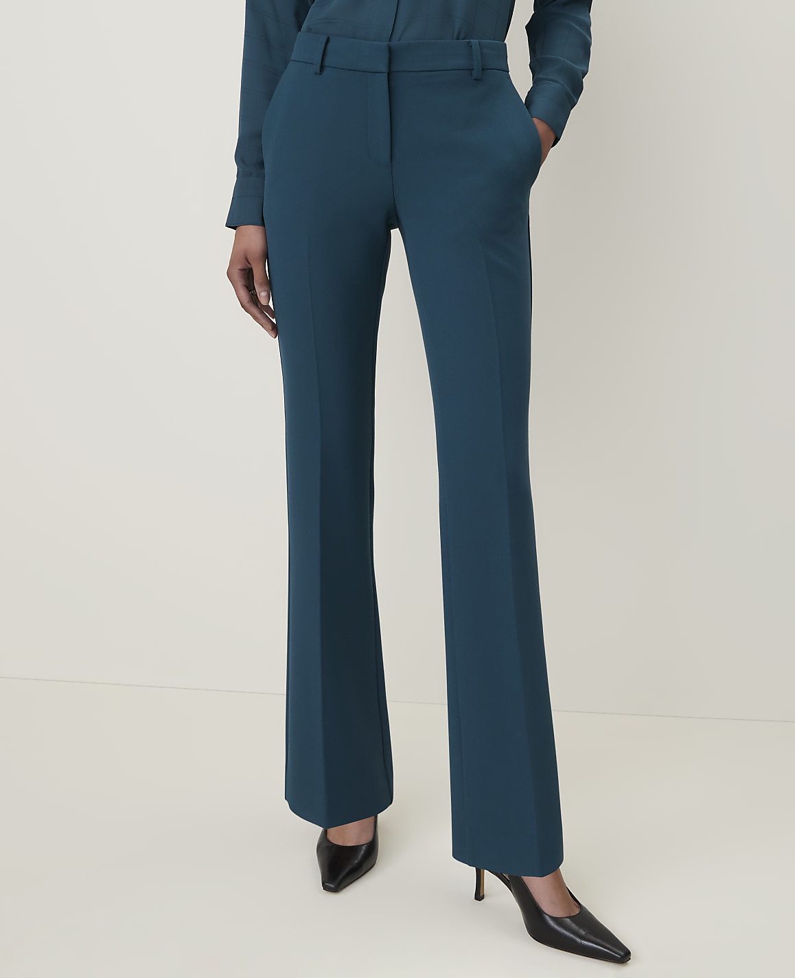A person stands in teal trousers and black heels against a plain, light background.